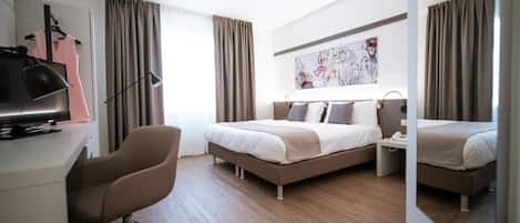 A modern hotel room with a bed, a desk with a computer, a chair, and a large mirror.