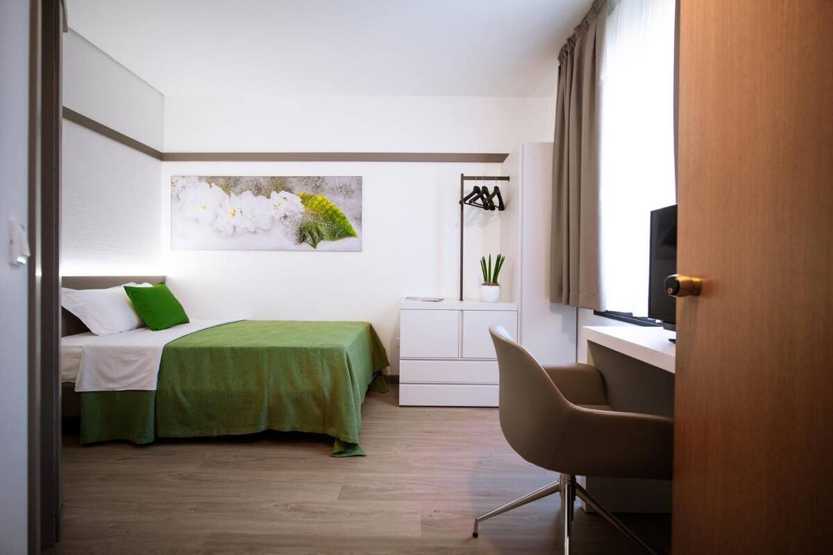 A bedroom with a bed, a desk, a chair, a television, and a wall-mounted plant.