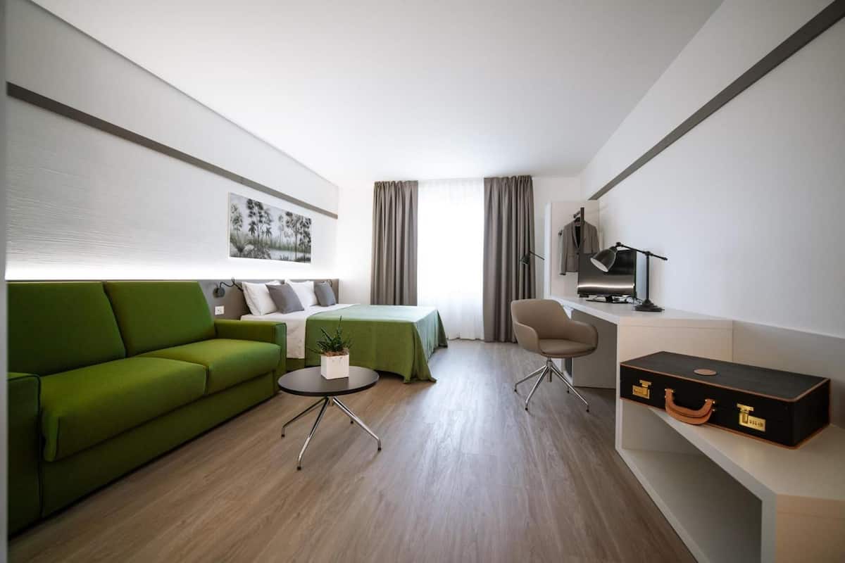 A modern hotel room with a green sofa, a bed, a desk with a lamp, and a suitcase.