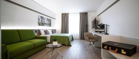 A modern hotel room with a green sofa, a bed, a desk with a lamp, and a suitcase.