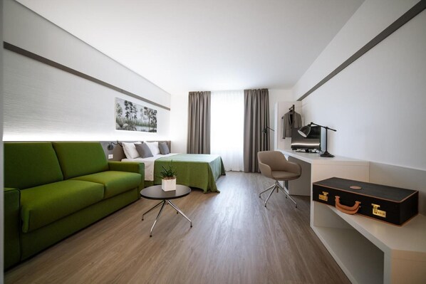A modern hotel room with a green sofa, a bed, a desk with a lamp, and a suitcase.