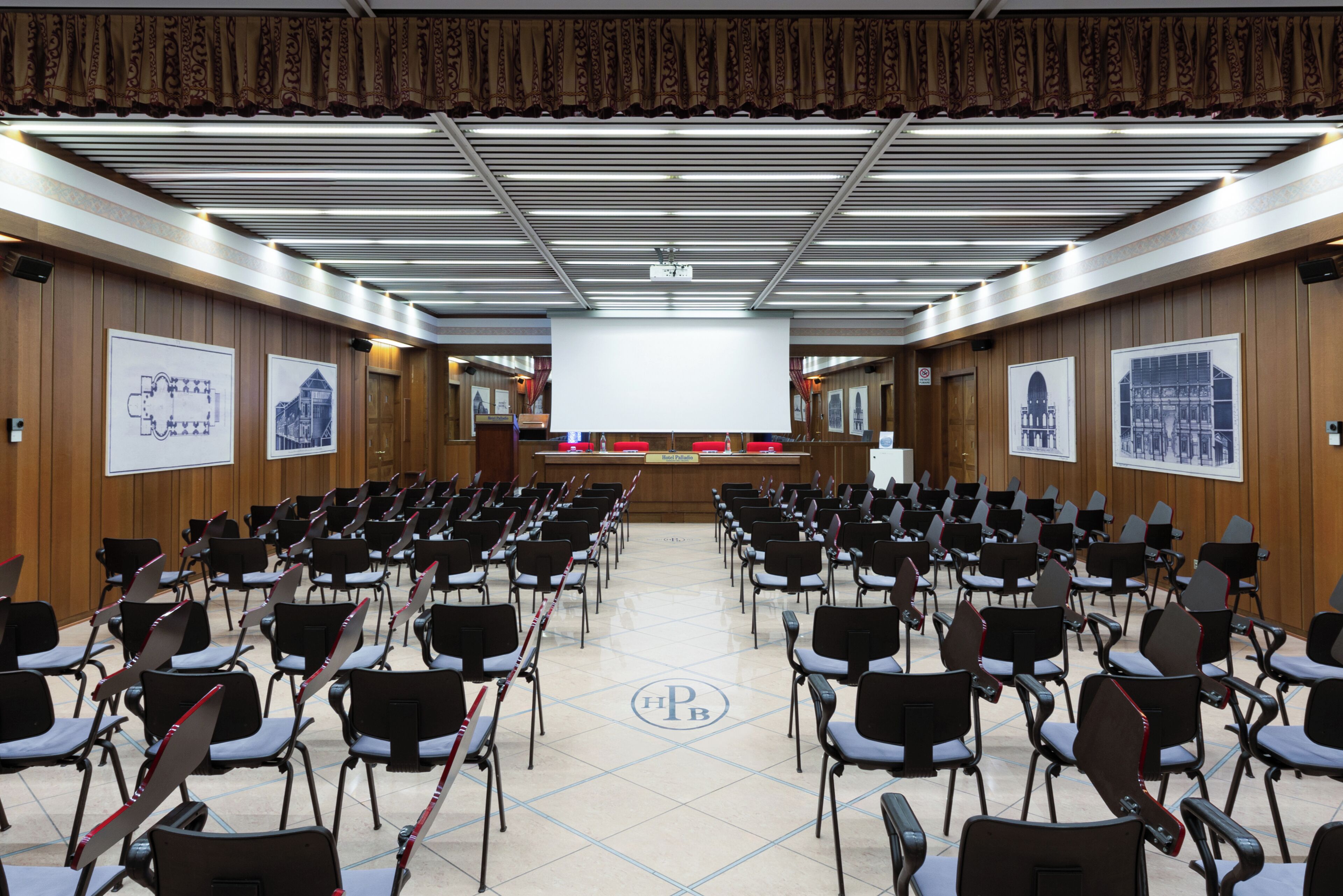 Un salón de conferencias con filas de sillas frente a un atril, paneles de madera y planos arquitectónicos enmarcados en las paredes.