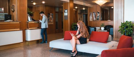 A reception area with a receptionist, a waiting area with a sofa and red cushions, and a reception desk with a computer and a plant.