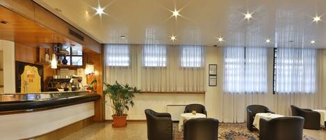 Un vestíbulo de hotel con mostrador de recepción, zona de estar y ventanas amplias con cortinas.