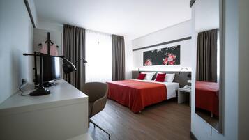 Habitación de hotel con cama, escritorio, silla, mesita de noche, espejo y ventana con cortinas.