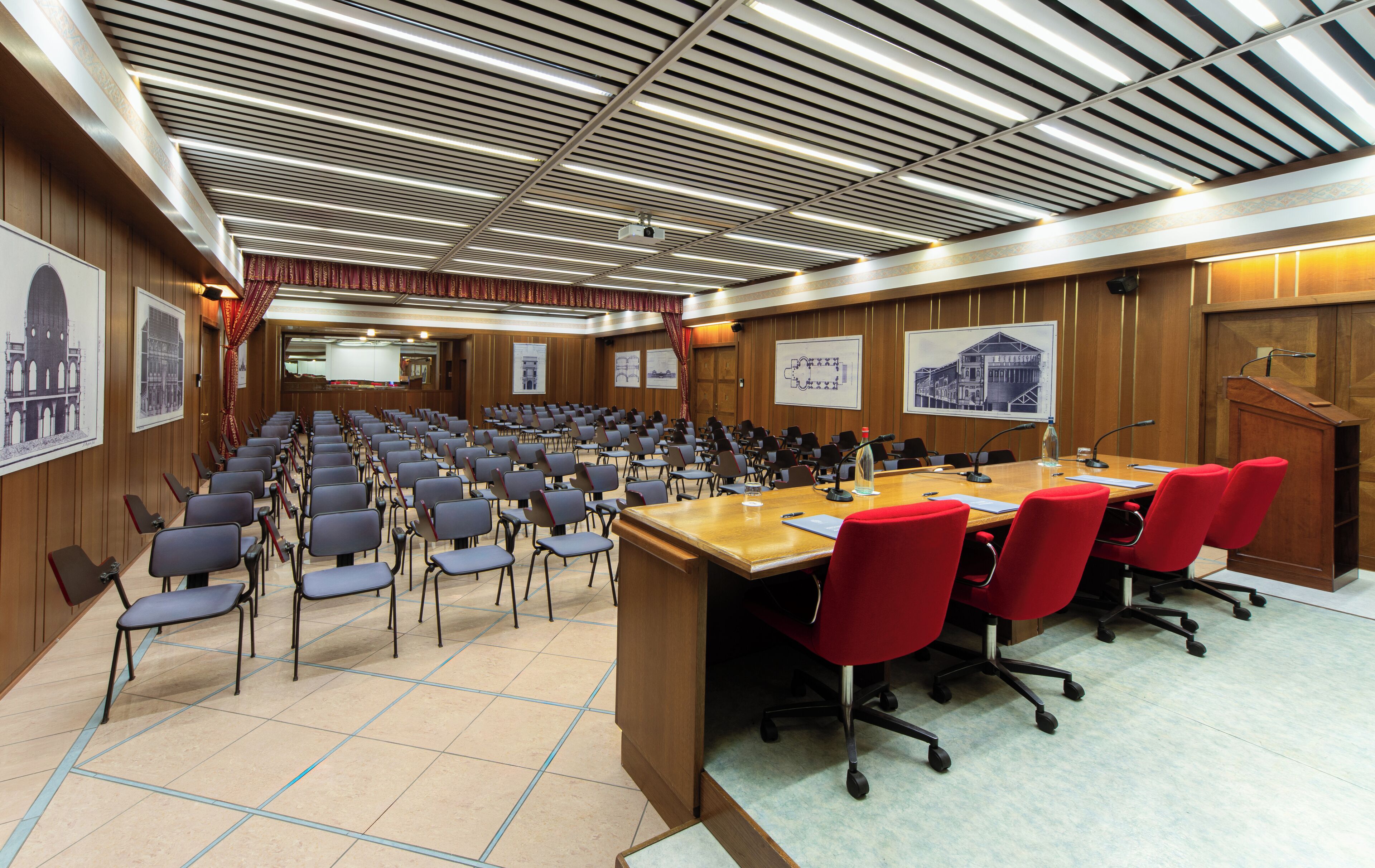 A conference room with rows of chairs, a long table with microphones, and a projector screen.