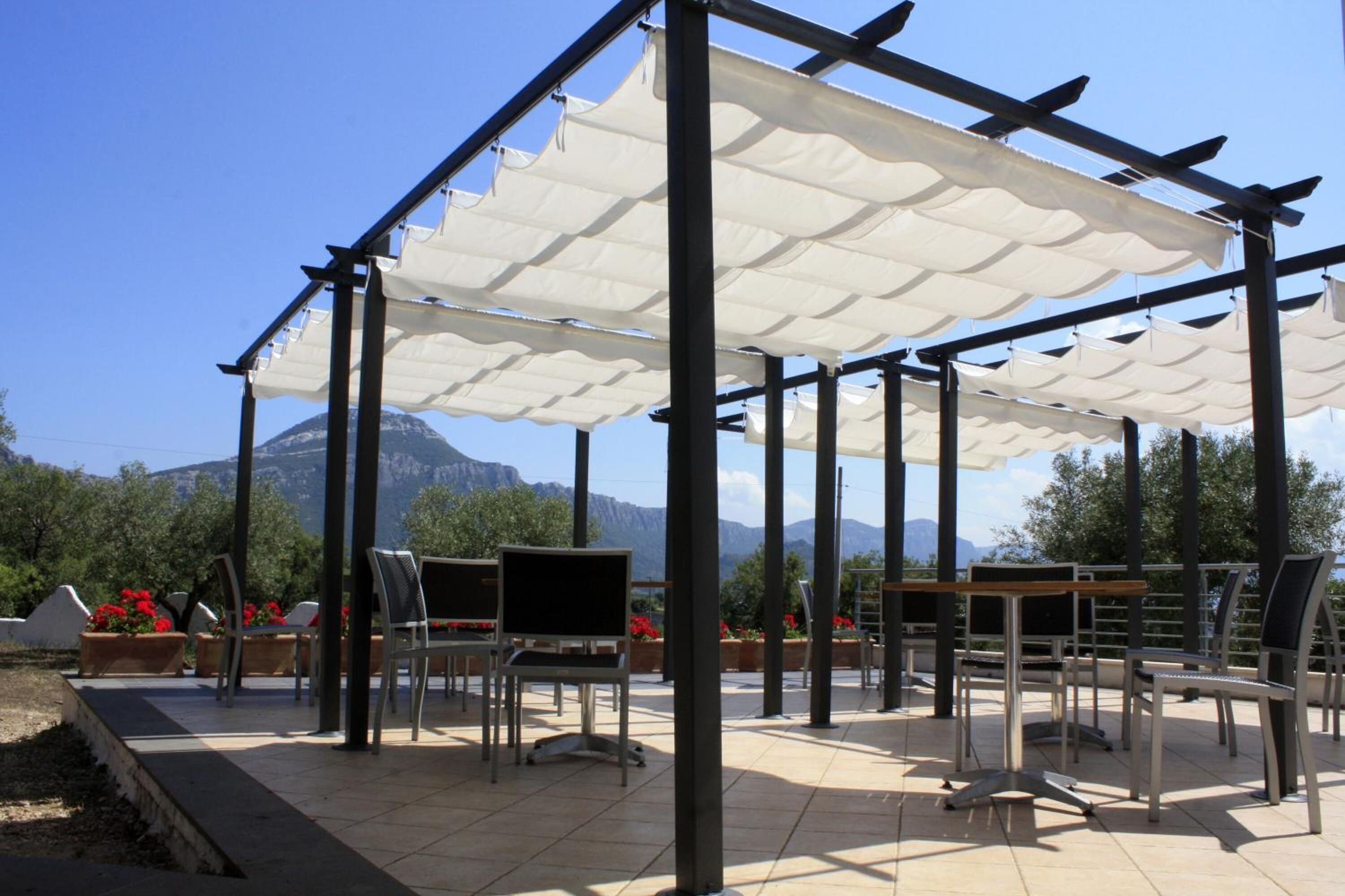 Un patio exterior con sillas y mesas de metal, toldos blancos y montañas al fondo.