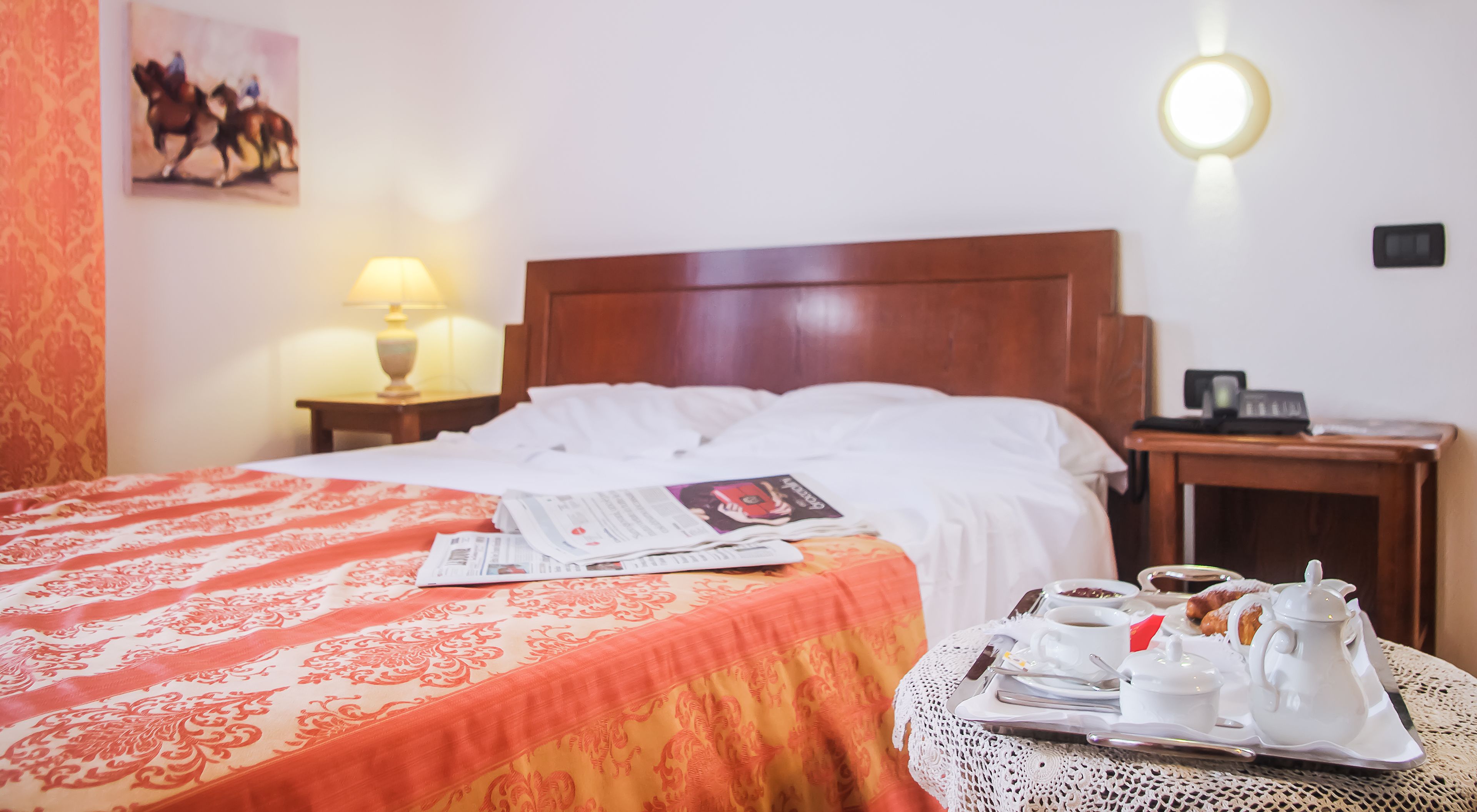 A hotel room with a neatly made bed, a bedside table with a lamp, a small table with breakfast items, and a painting on the wall.