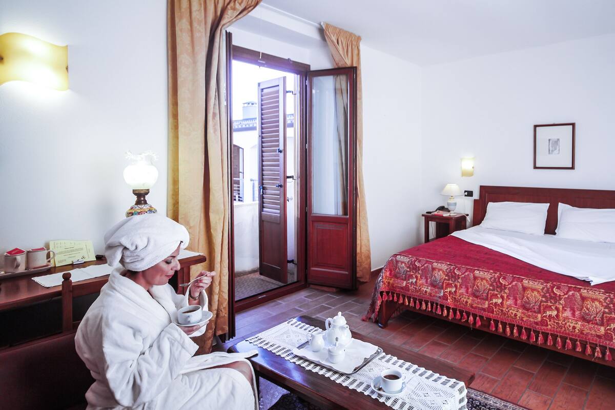 A hotel room with a bed, a bedside table with a lamp, a chair, and a table with a tray of tea and cups.