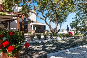 Un patio con una pérgola, geranios en macetas, una estatua y vistas al mar.