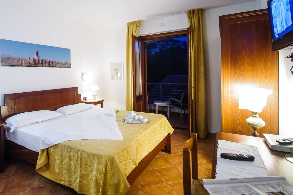 A hotel room with a bed, a bedside table with a lamp, a chair, a TV, and a window with curtains.