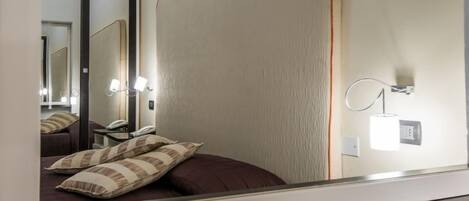 Una habitación de hotel con cama, mesita de noche, espejo y lámpara de pared.