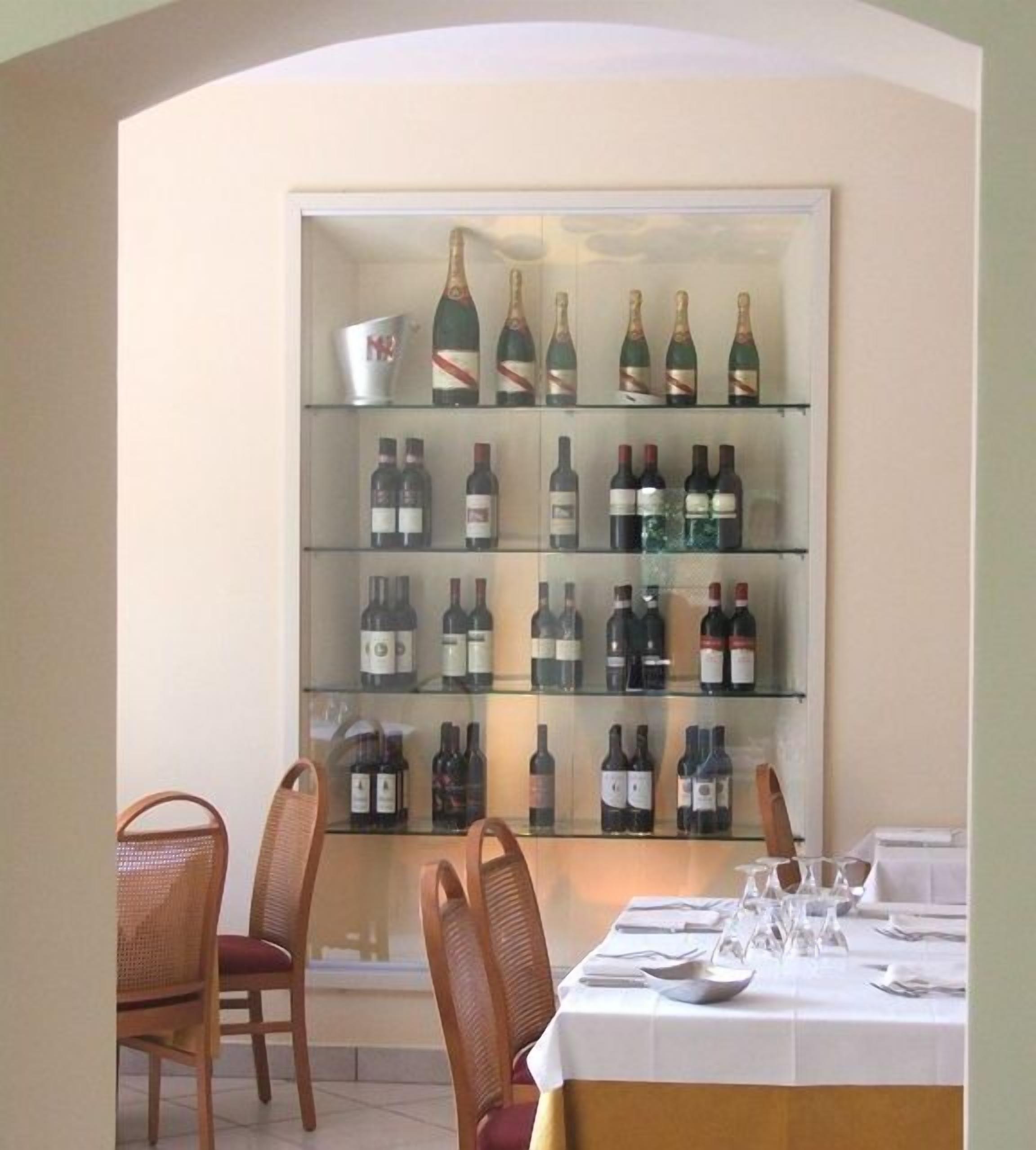 A dining area with a long table set for a meal, surrounded by wicker chairs. Behind the table, there is a wall-mounted wine rack with multiple shelves holding various bottles of wine.