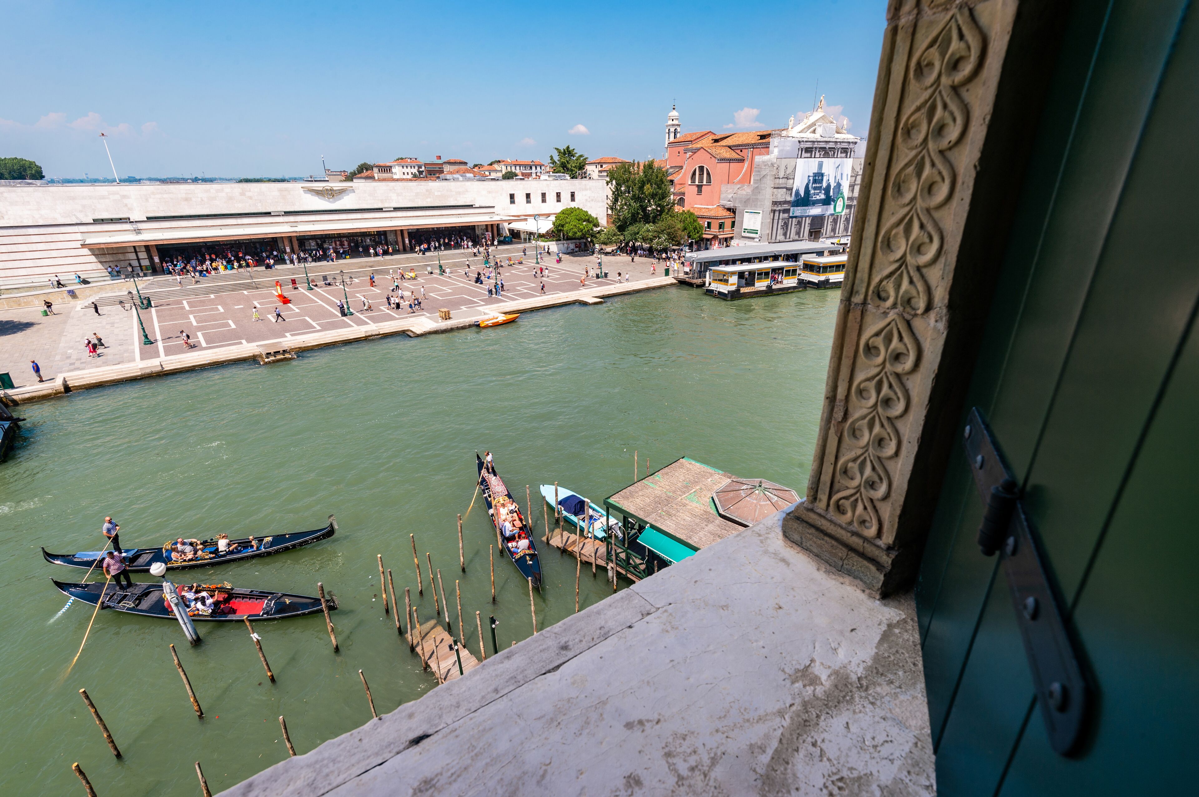 Une vue d’un canal où des gondoles sont amarrées au quai, d’un grand bâtiment surmonté d’un dôme, et d’une foule de personnes en arrière-plan.