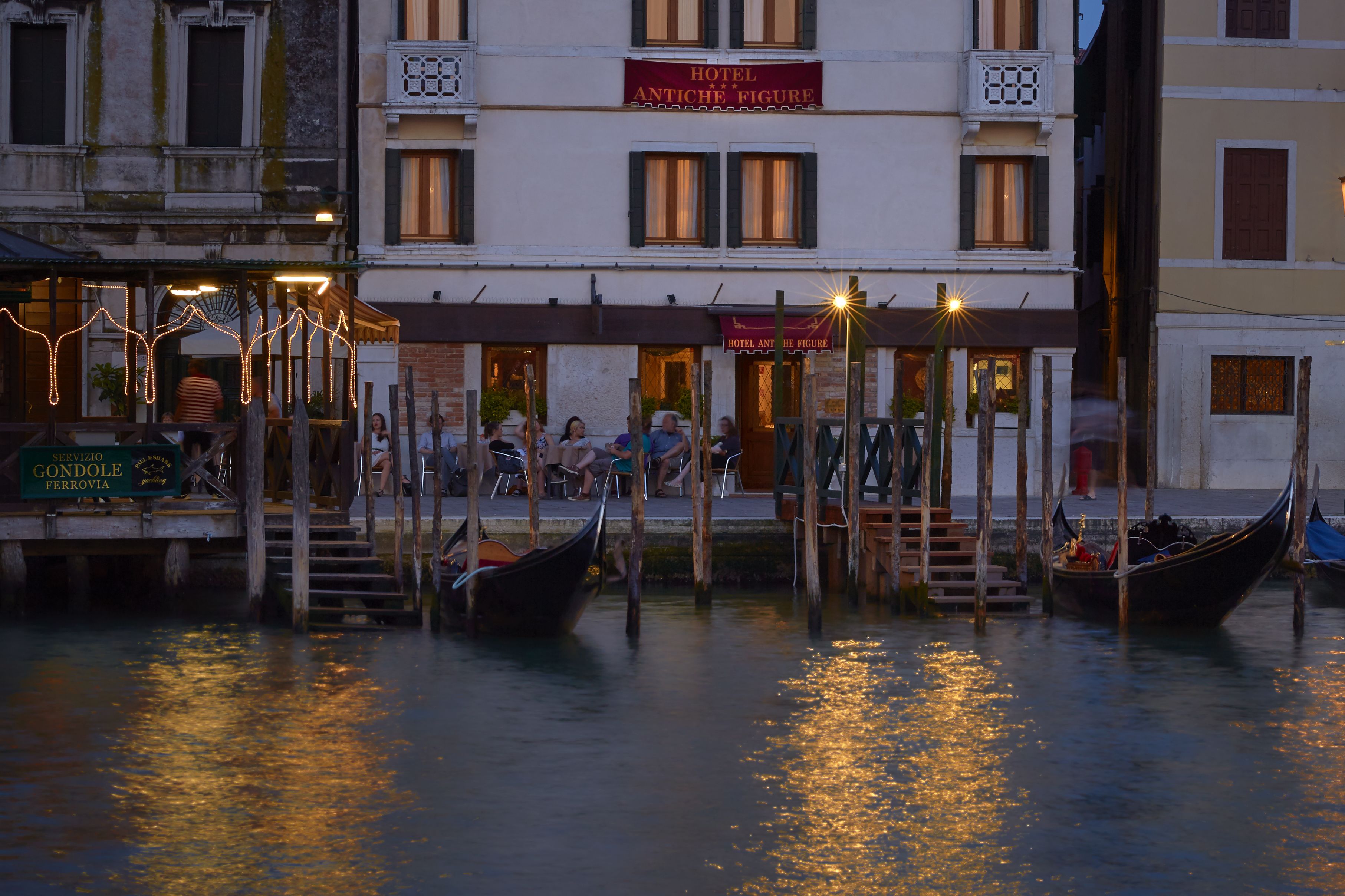 Un hotel sul lungomare con gondole ormeggiate nell'acqua, una terrazza con posti a sedere all'aperto e un cartello che recita "HOTEL ANTICHE FIGURE".