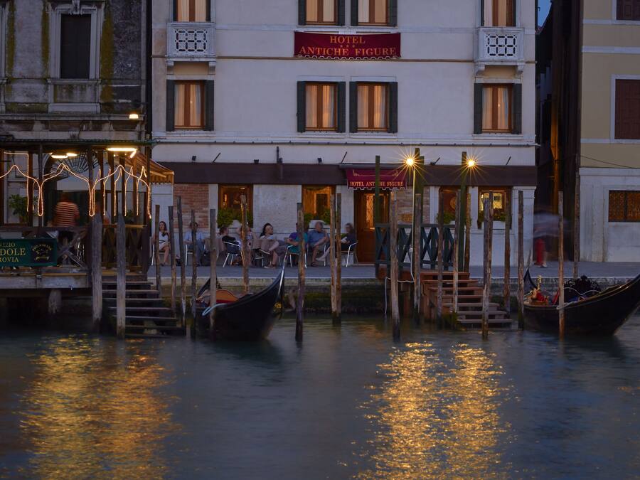 Un hotel sul lungomare con gondole ormeggiate nell'acqua, una terrazza con posti a sedere all'aperto e un cartello che recita "HOTEL ANTICHE FIGURE".