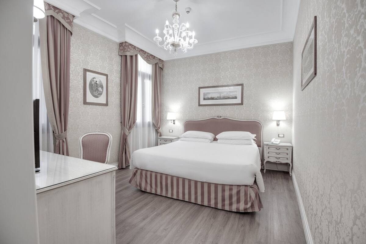 Ein Hotelzimmer mit einem großen Bett, zwei Nachttischen, einem Sessel, einer Chandelüster und einem gerahmten Bild an der Wand.