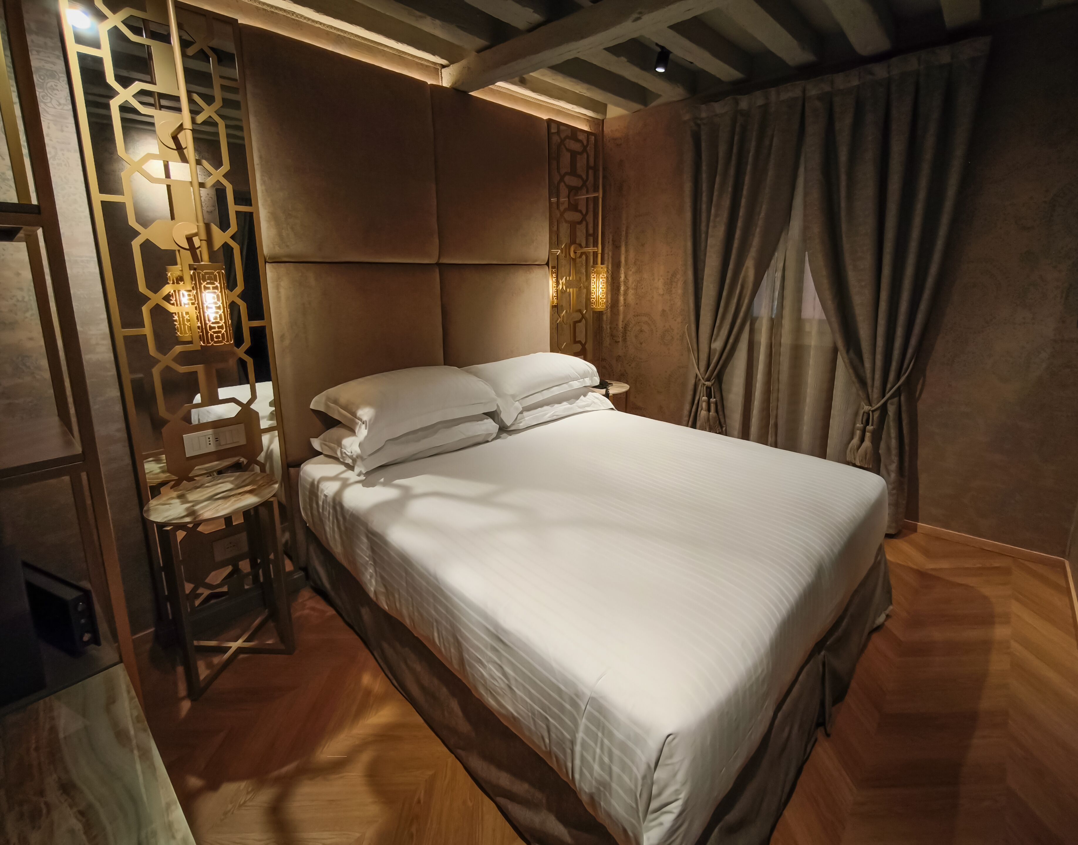 A neatly made bed in a room with a wooden floor, a decorative wall panel, and a curtain on the window.