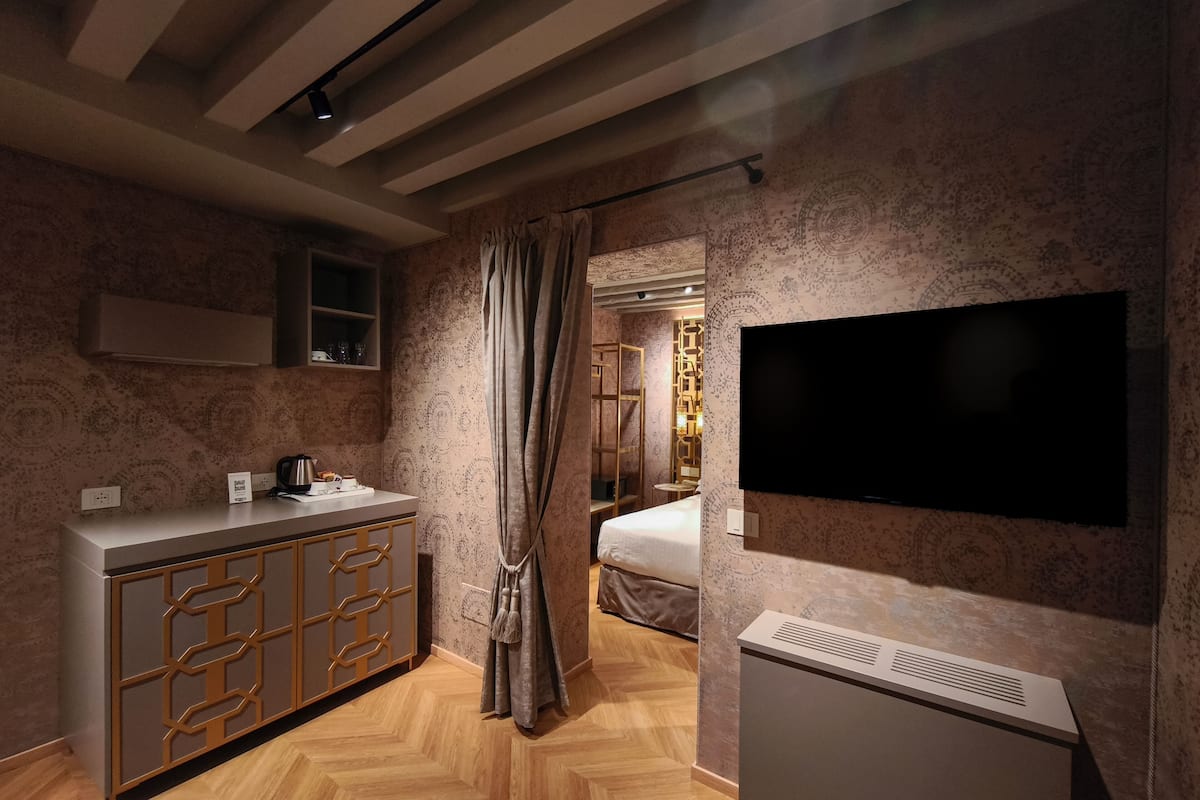 Ein modernes Hotelzimmer mit einem großen Flachbildfernseher an der Wand, einem Holzboden im Fischgrätmuster, einer integrierten Kommode mit geometrischem Design und einem Bett mit Kopfteil.