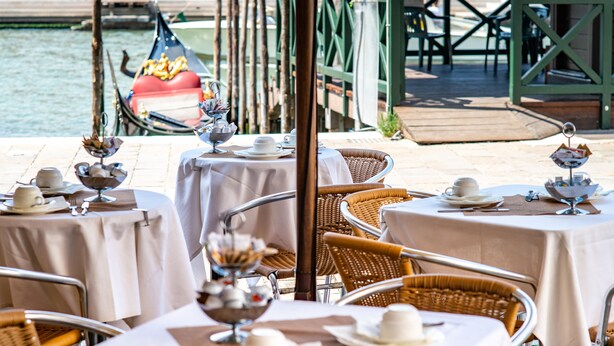 Un espace de restauration en plein air avec des tables dressées pour un repas, des chaises en osier et une gondole en arrière-plan.