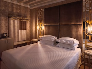 Quarto de hotel com uma cama grande, uma mesa de cabeceira com chaleira e arandelas decorativas.