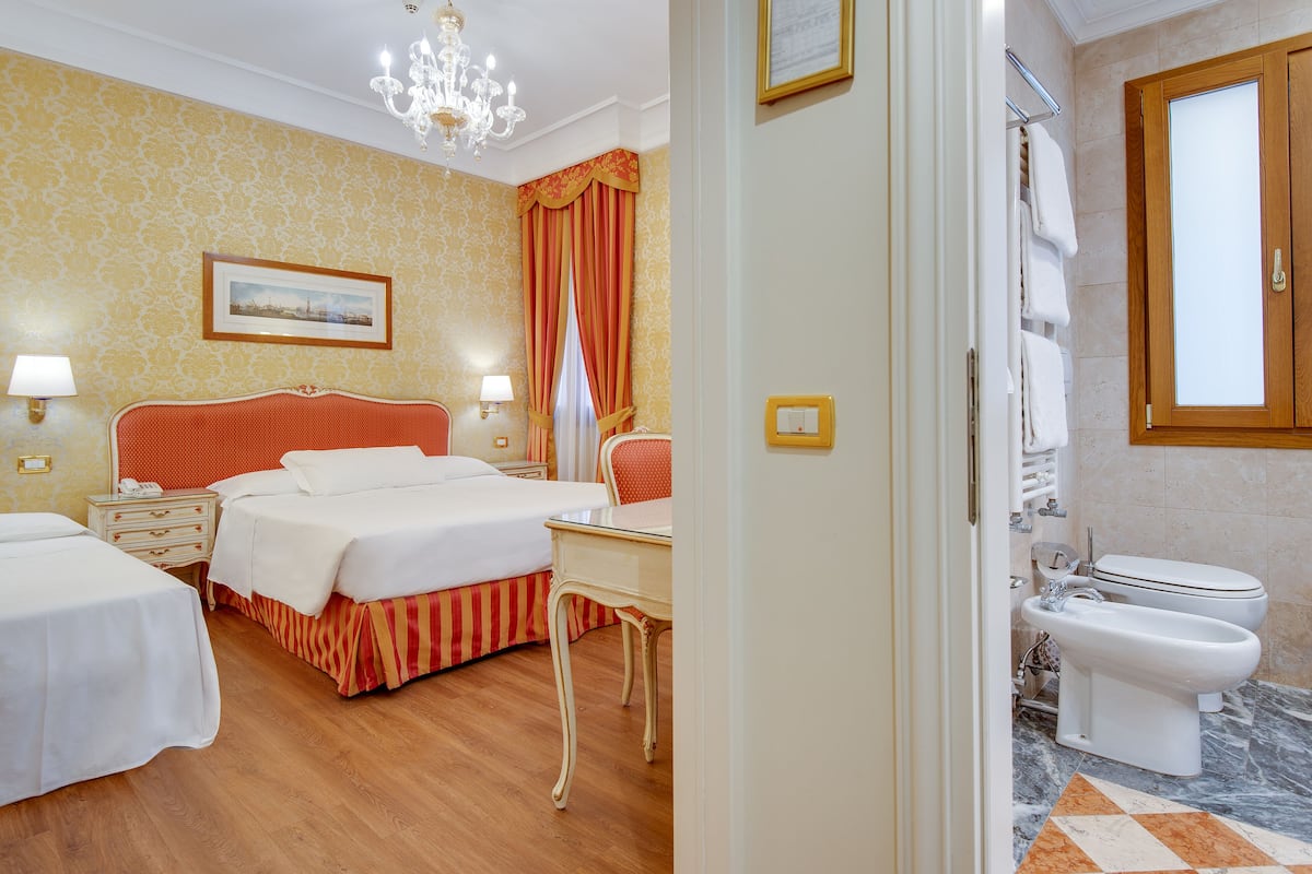 Ein Hotelzimmer mit zwei Betten, einem Schreibtisch, einer Chandelierleuchtre und einem Badezimmer mit Toilette und Bidet, sowie einer Wand mit Muster.
