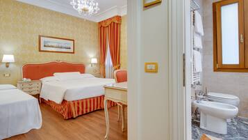 Une chambre d’hôtel avec deux lits, un bureau, un lustre, une salle de bain avec des toilettes et un bidet, et un mur décoré de motifs.