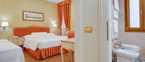 A hotel room with two beds, a desk, a chandelier, a bathroom with a toilet and bidet, and a patterned wall.