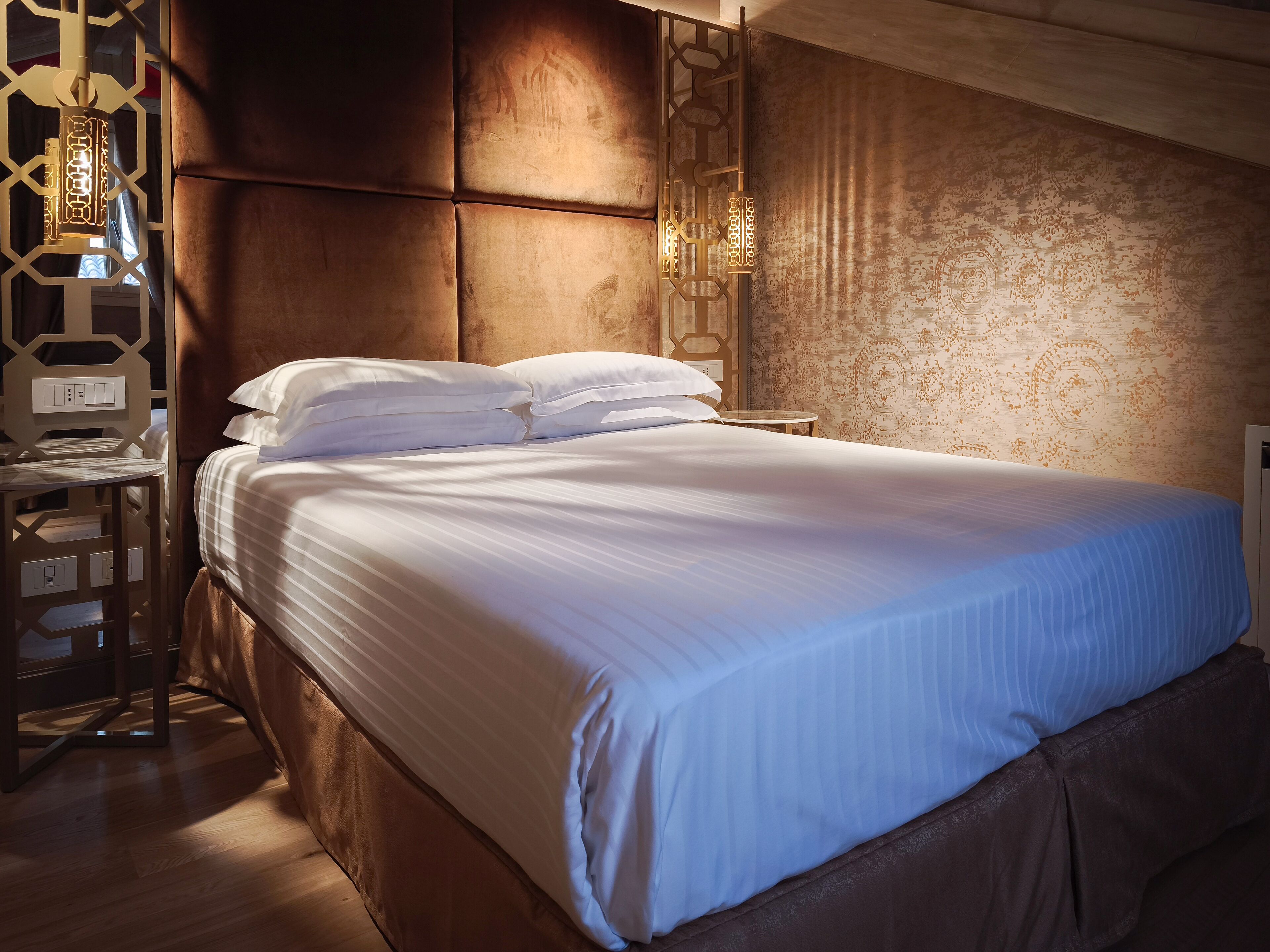 A neatly made bed with white linens, a wooden headboard, and a patterned wall behind it.