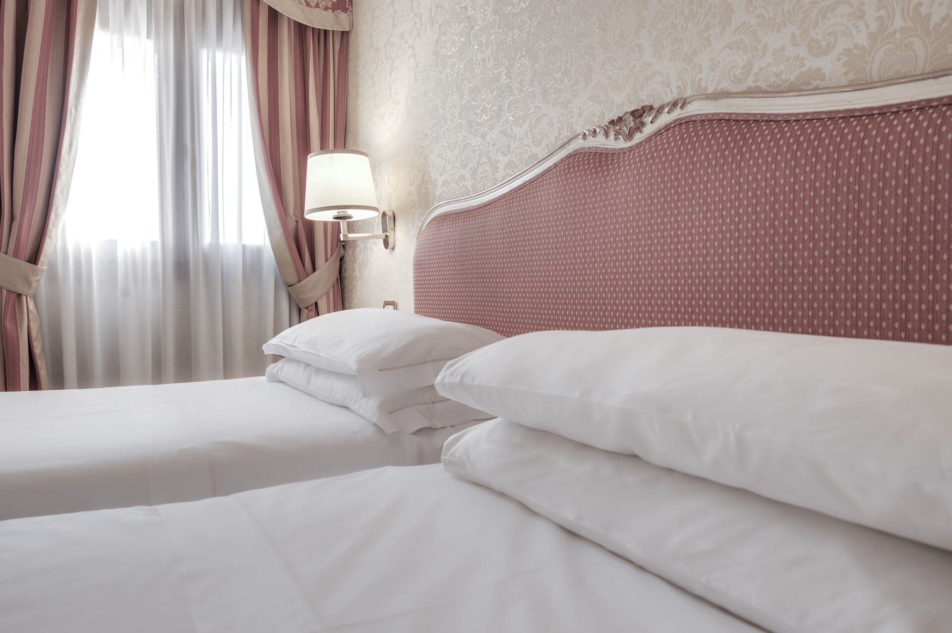 A hotel room with a large bed, two pillows, a nightstand with a lamp, and a window with curtains.