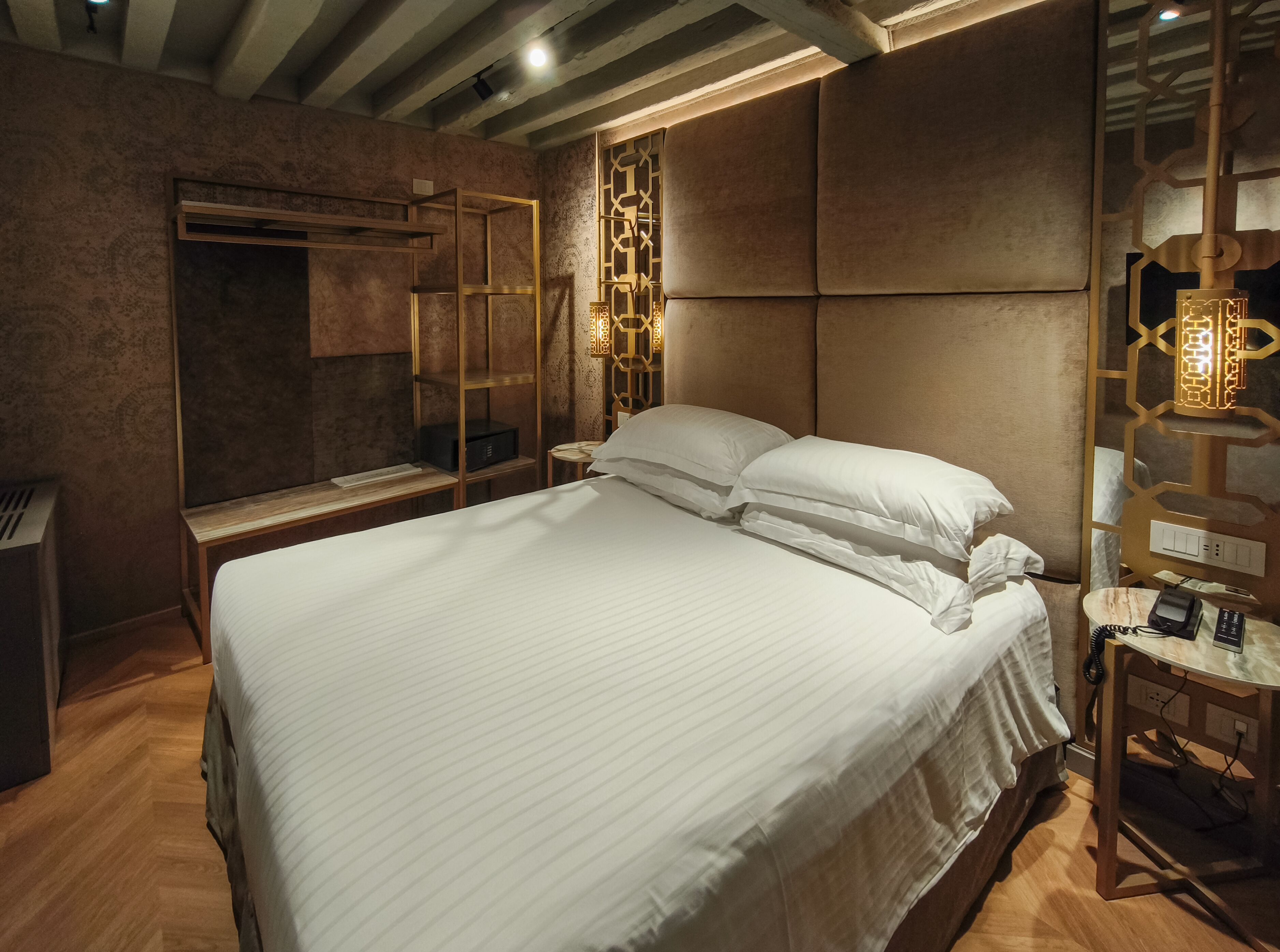 A modern hotel room with a large bed, a wooden bench, a vanity with a mirror, and a decorative wall panel.