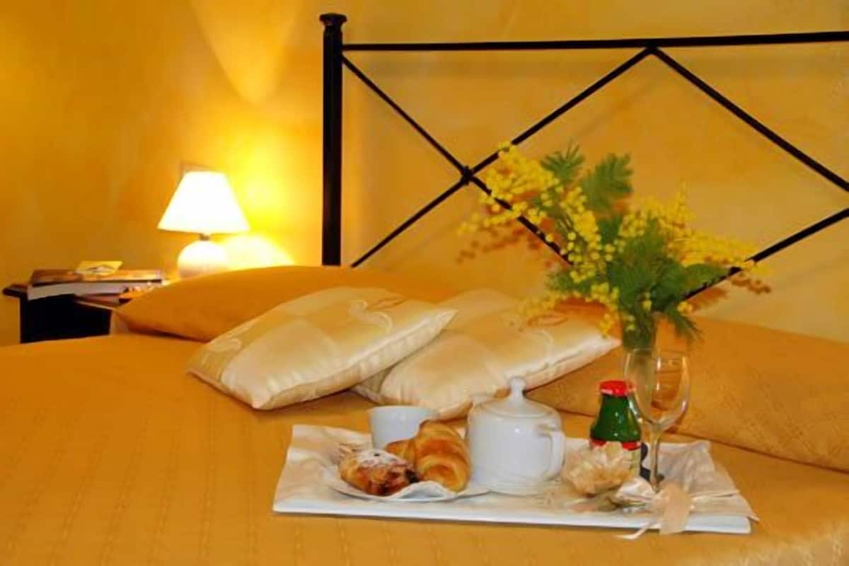 Un lit avec une tête de lit jaune, des oreillers, une table de chevet avec une lampe, et un plateau avec des croissants, une bouteille et un verre.