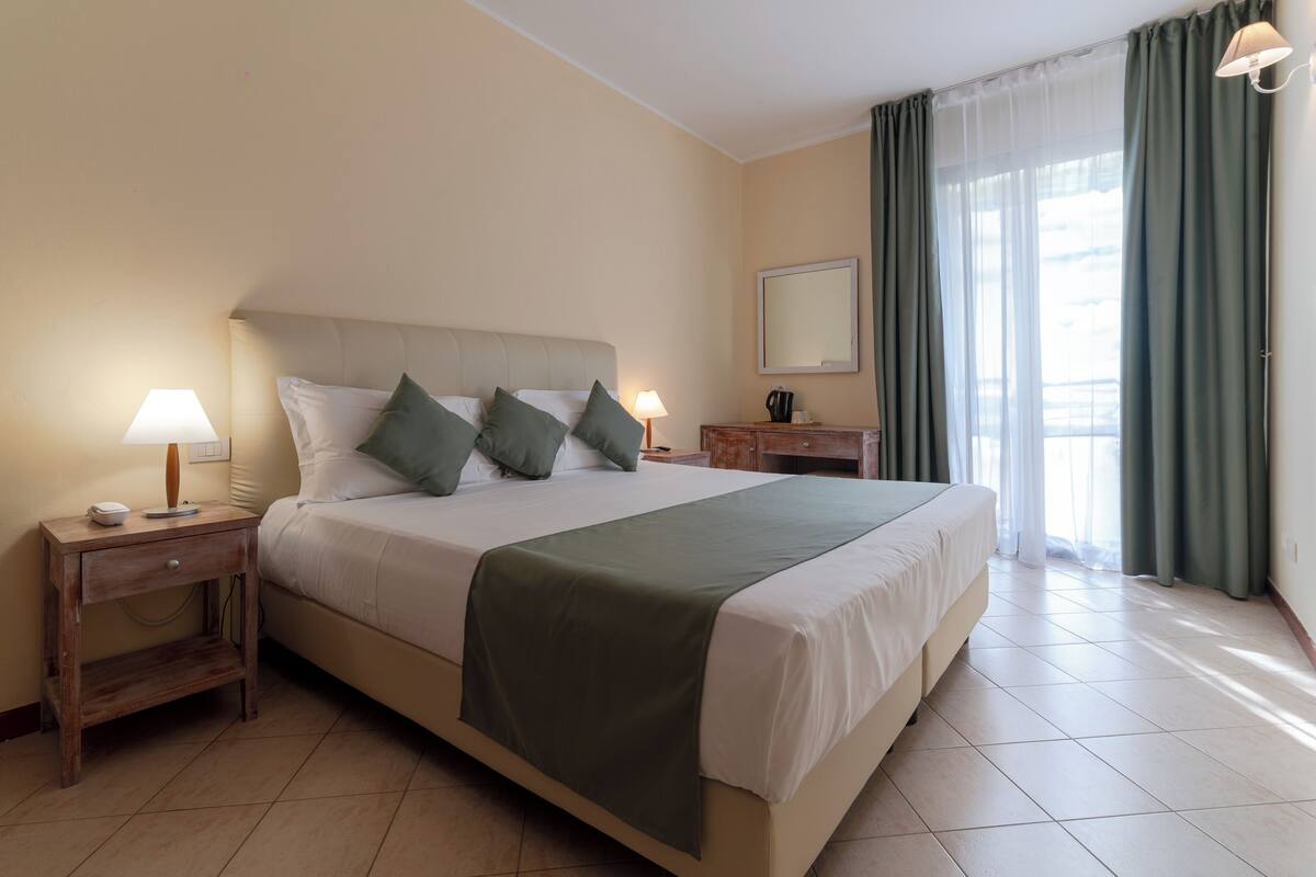 A hotel room with a large bed, two bedside tables with lamps, a bedside mirror, and a window with curtains.