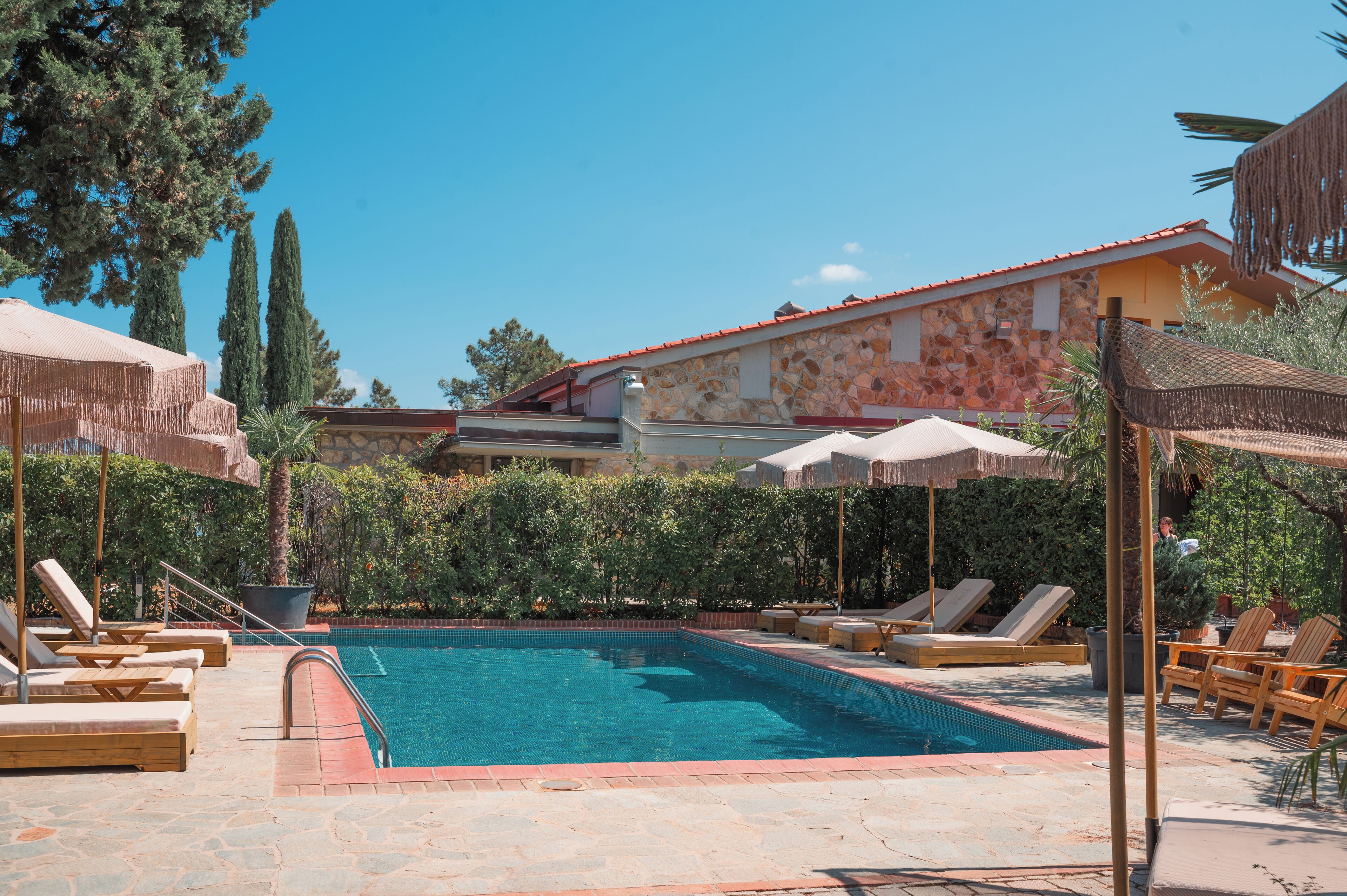A swimming pool with lounge chairs and umbrellas, surrounded by greenery and a stone building.