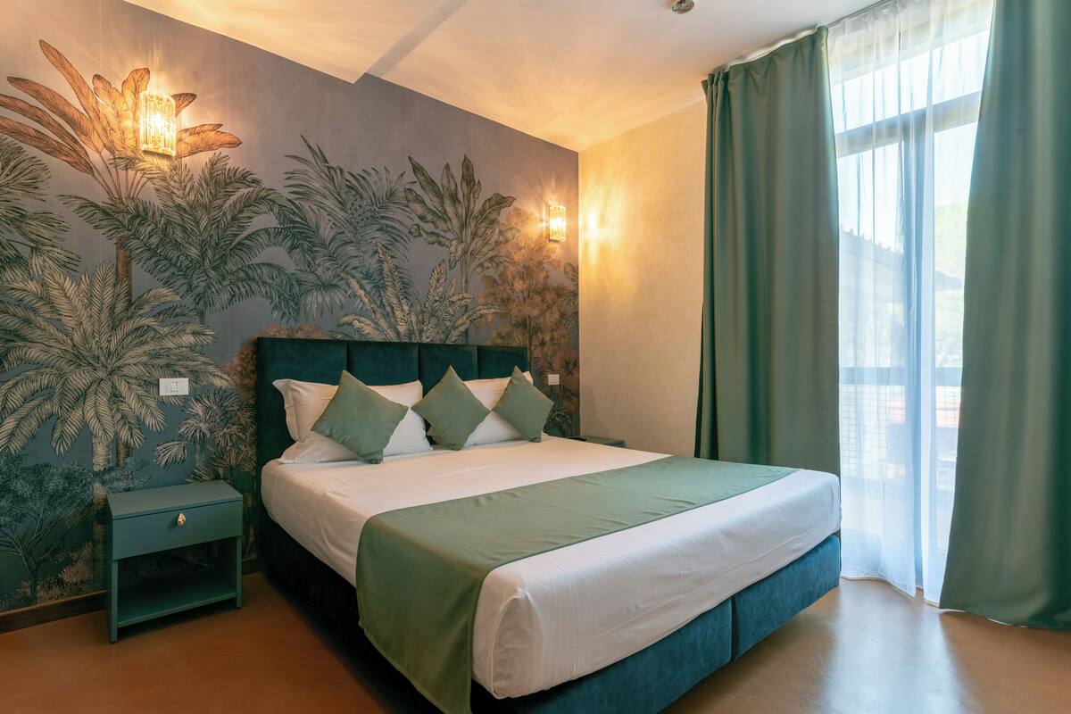 A neatly made bed with a green bedspread and white pillows, a nightstand with a drawer, a wall with a palm tree mural, and a window with green curtains.