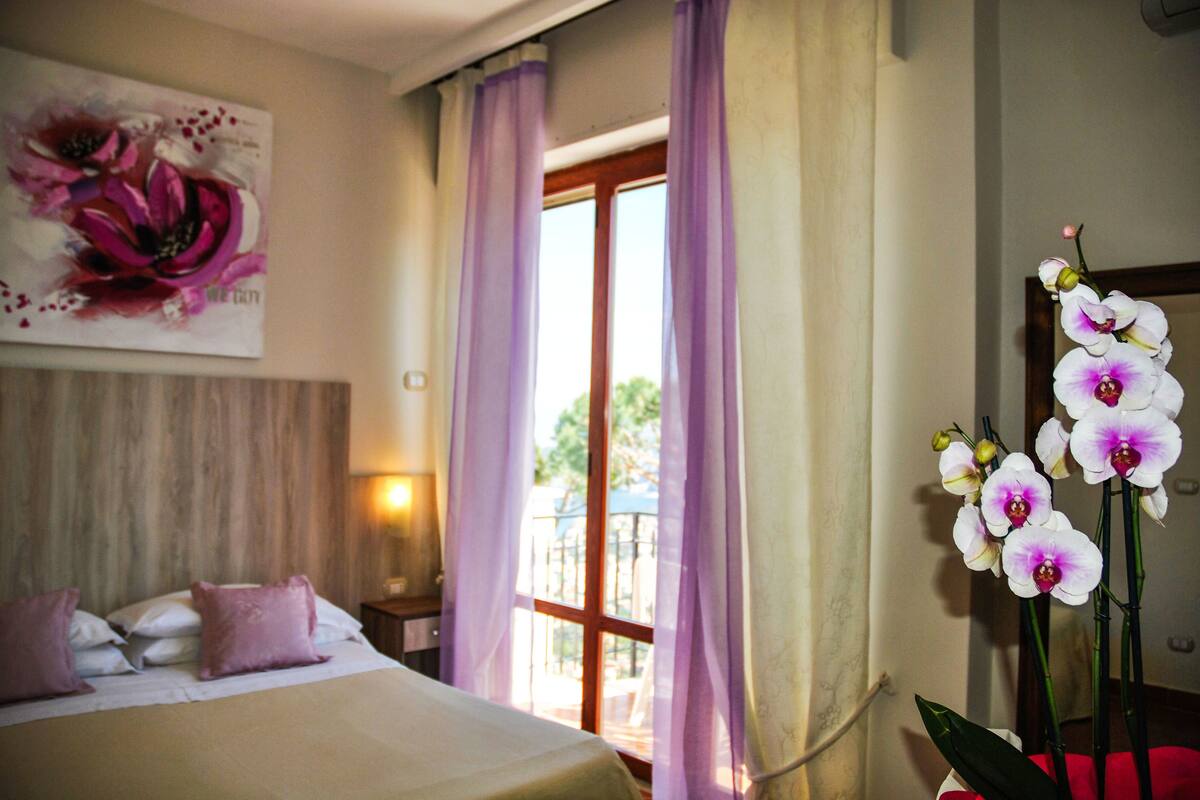 A hotel room with a bed, a window with purple curtains, and a large floral painting on the wall.
