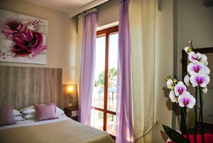 A hotel room with a bed, a window with purple curtains, and a large floral painting on the wall.