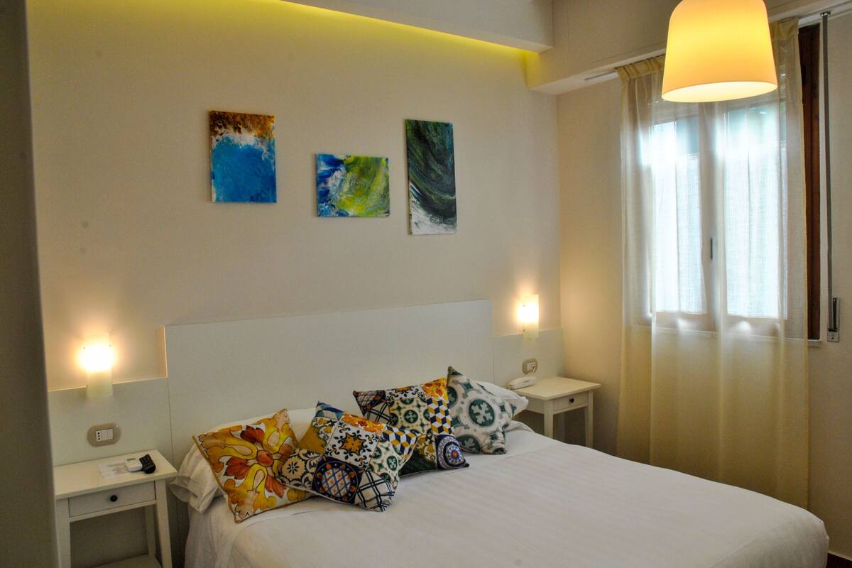 A hotel room with a bed, two bedside tables, a nightstand, a lamp, and artwork on the wall.