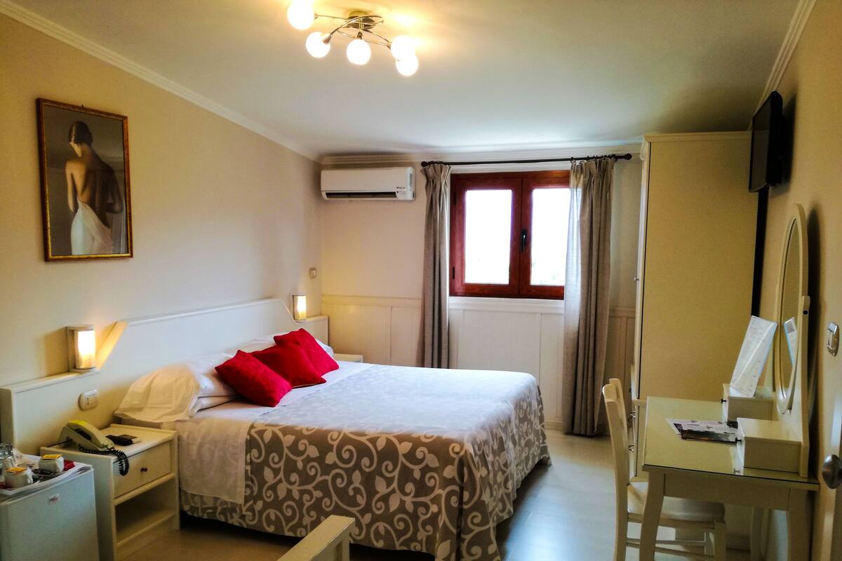 A hotel room with a bed, bedside table with a lamp, a desk with a chair, an air conditioning unit, and a painting on the wall.
