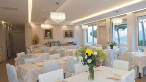 A dining area with round tables, white tablecloths, and chairs. There are chandeliers hanging from the ceiling, and large windows with curtains. - Hotel Villa Fiorita (Sorrento)