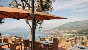 An outdoor terrace with wicker furniture and a large umbrella, overlooking a coastal landscape with mountains and a city in the distance. - Hotel Villa Fiorita (Sorrento)