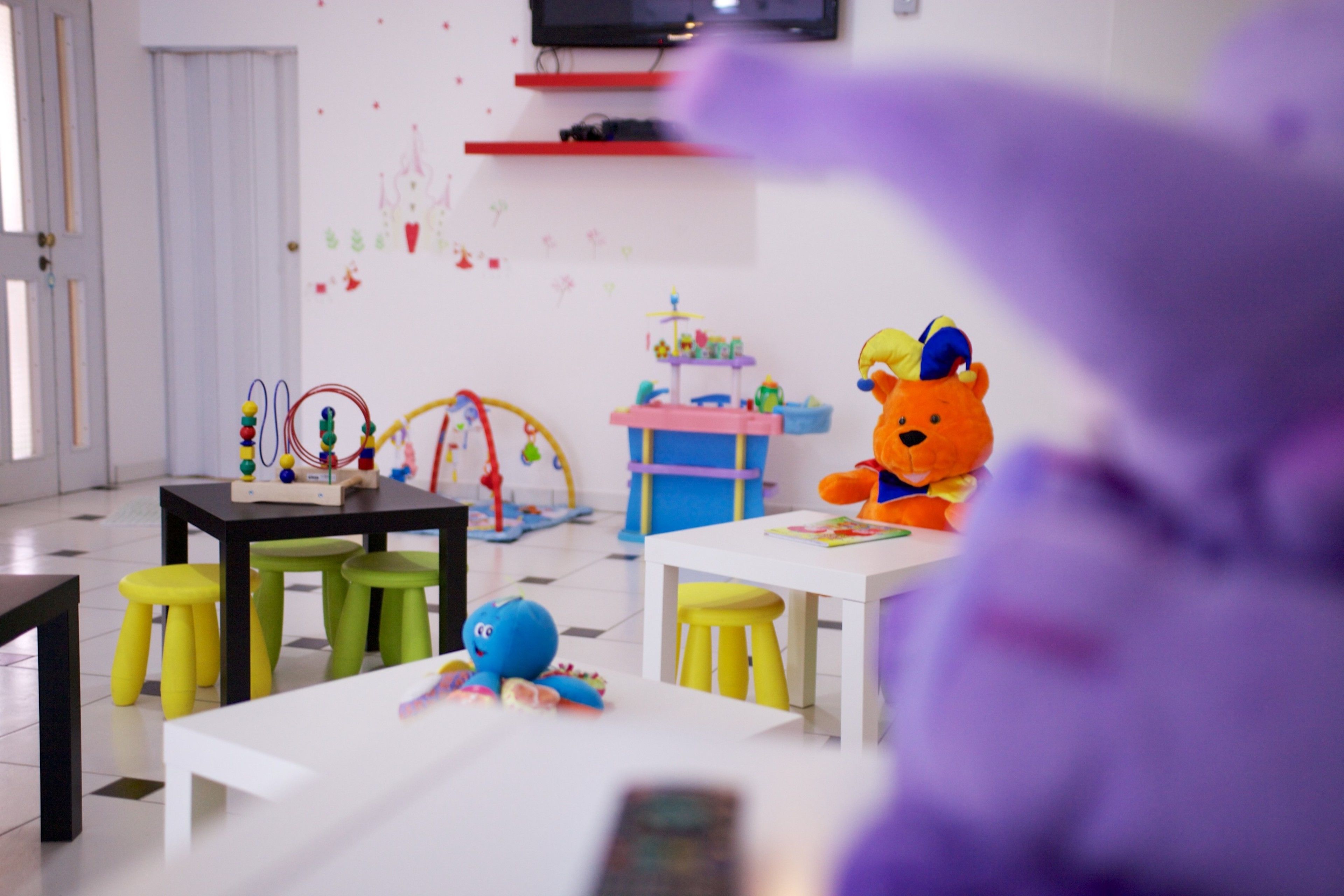 A children's playroom with a white table, green stools, a teddy bear, and a colorful play area.