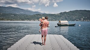 A pier with a boat in the background, a person holding a child, and mountains in the distance. - Hotel Carillon (Baveno)