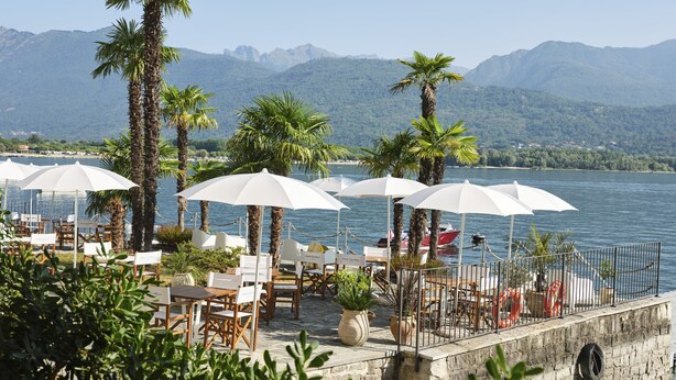 A lakeside restaurant with outdoor seating, palm trees, and a view of mountains. - Hotel Carillon (Baveno)