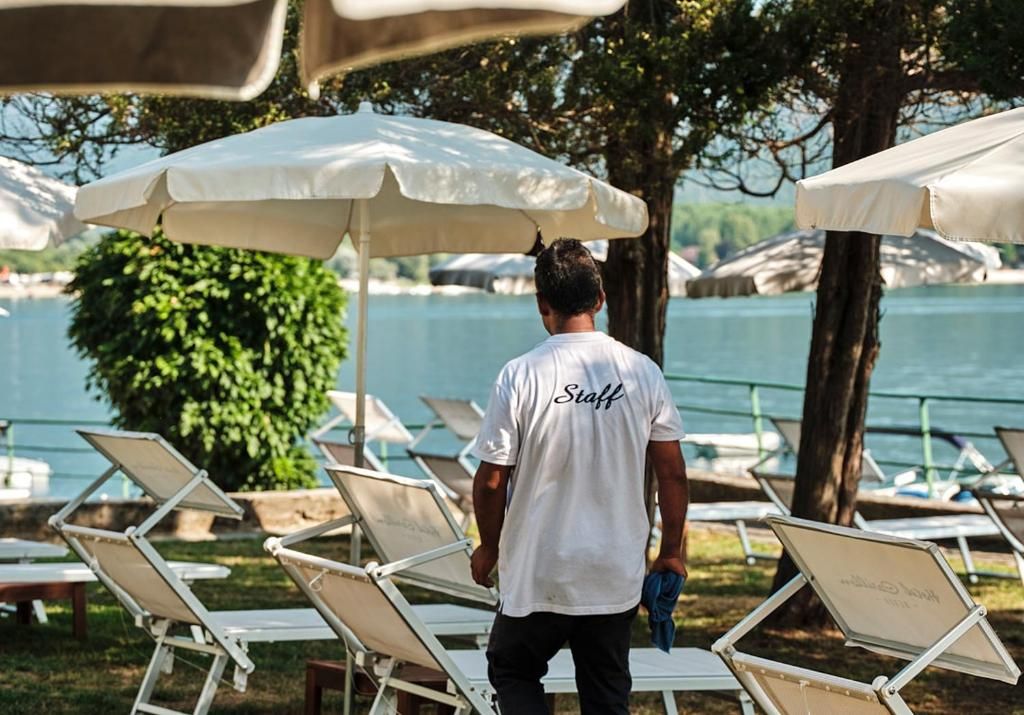 A person in a white shirt with 'Staff' on the back walking past sun loungers and umbrellas by a body of water.