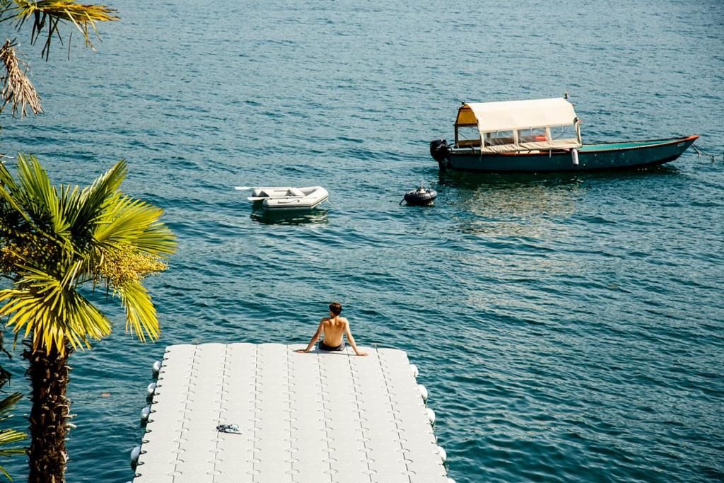 Una persona sentada en un muelle, con un bote y una embarcación más pequeña en el agua.