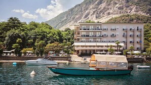 A coastal hotel with a mountain backdrop, a marina with boats, and outdoor seating. - Hotel Carillon (Baveno)