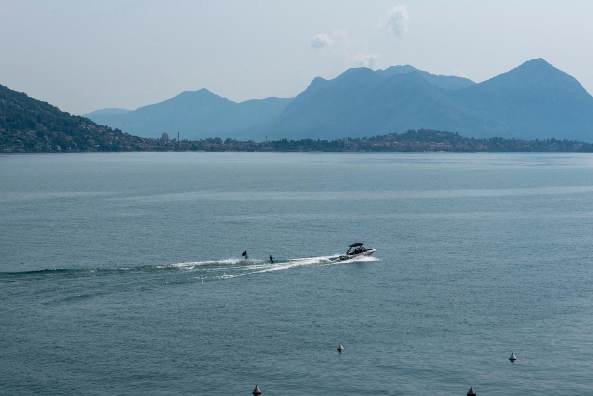 Un cuerpo de agua con un bote y un jet ski, montañas al fondo y una costa lejana con edificios.