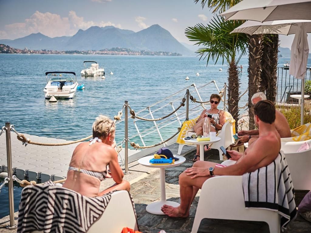 Un gruppo di persone sedute a tavoli vicino al lungomare, con barche sullo sfondo e montagne in lontananza.