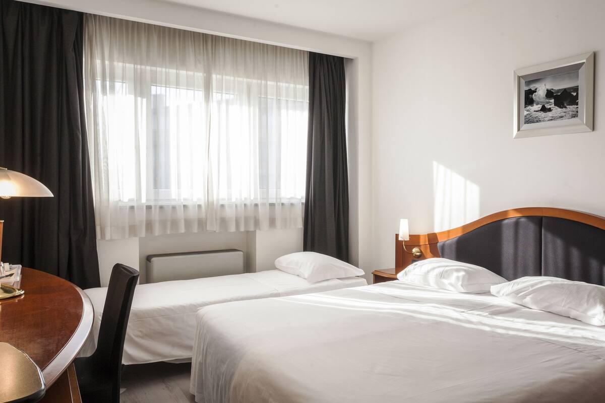 A hotel room with two single beds, a wooden desk, a chair, a lamp, a framed picture on the wall, and a window with sheer curtains.